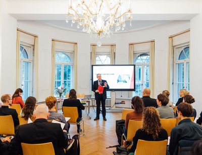 Das Bild zeigt die Kick-Off Veranstaltung zum Pre-Doc Award. Auf dem Bild ist eine vortragende Person sowie mehrere Personen, die auf Stühlen ringsum sitzen. 