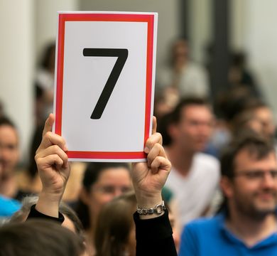 The picture shows a hand holding up a number: ‘7’ to judge the Science Slam.